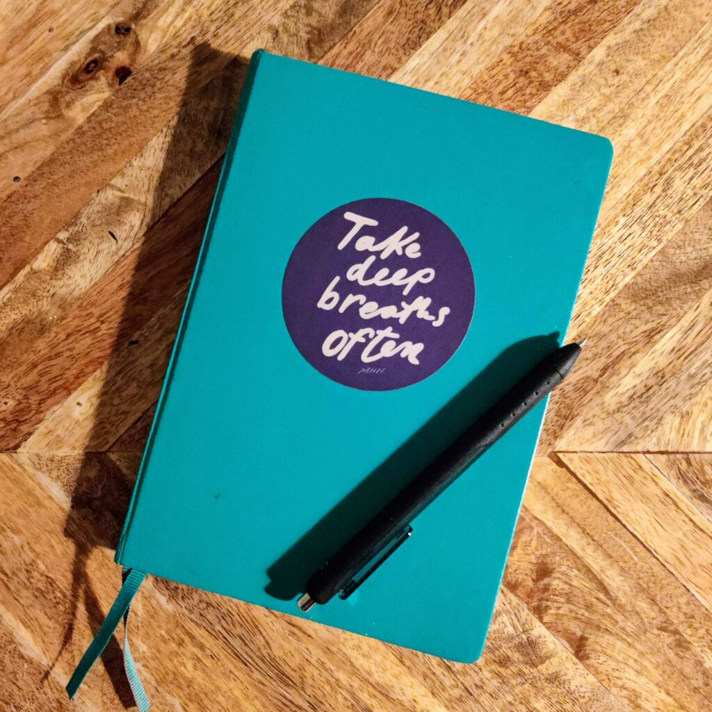 A teal book on a wooden table. There's a deep blue sticker that says "Take deep breaths often - MHN" and a black pen on top of the book.