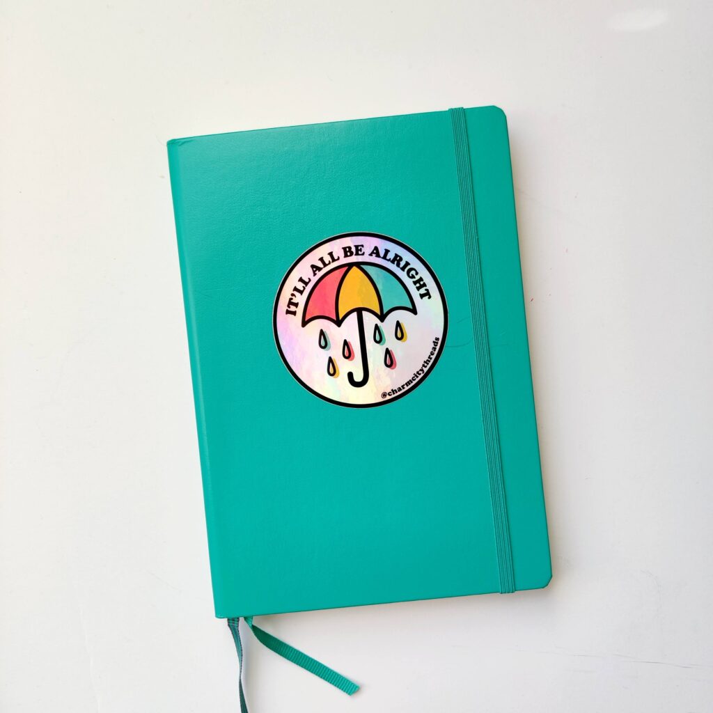 A turquoise Leuchtturm notebook with a Charm City Threads holographic sticker of an umbrella with the words "It'll be alright".