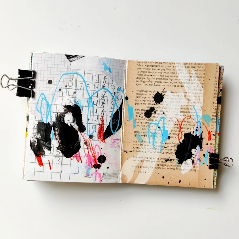 An open mixed media art journal. The pages are covered in a layer of collage, and on top of that are energetic marks in light blue, black, pink, white, and red. Most of the background peeks through.
