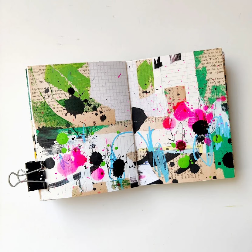 An open mixed media art journal. The pages are covered in a layer of collage, and on top of that are energetic marks in hot pink, light blue, green, and black. Most of the background peeks through.