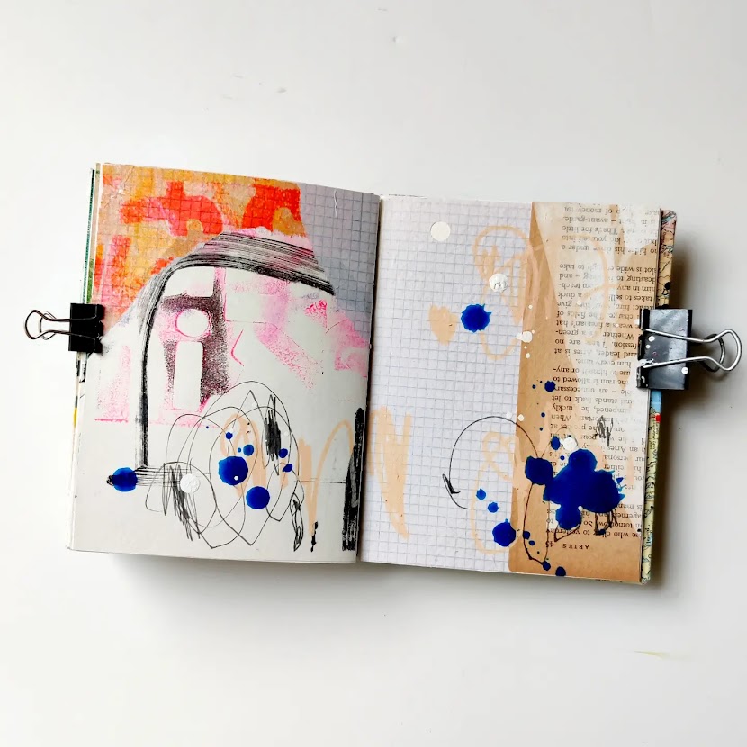 An open mixed media art journal. The pages are covered in a layer of collage, and on top of that are energetic marks in blue, black, and peach. Most of the background peeks through.