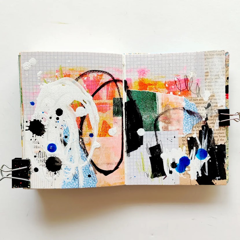 An open mixed media art journal. The pages are covered in a layer of collage, and on top of that are energetic marks in white, blue, and black. Most of the background peeks through.
