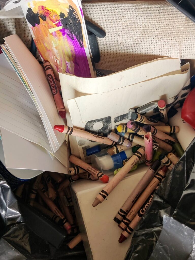 Close up of a trash bag containing various art supplies, including crayons, painted papers, charcoal, etc.