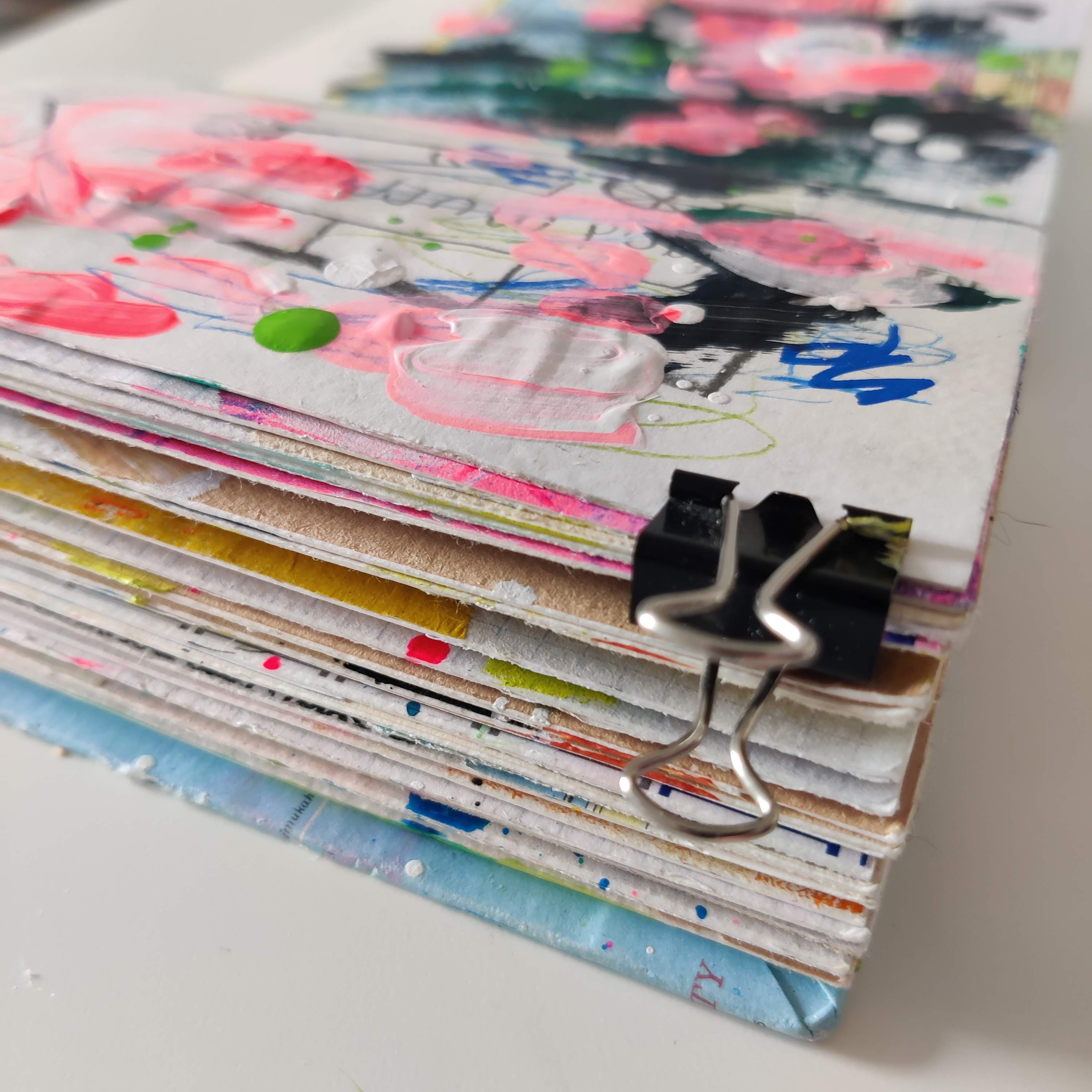 The corner of an open mixed media art journal. The page includes bright pink paint and elements of white, blue, and lime green.