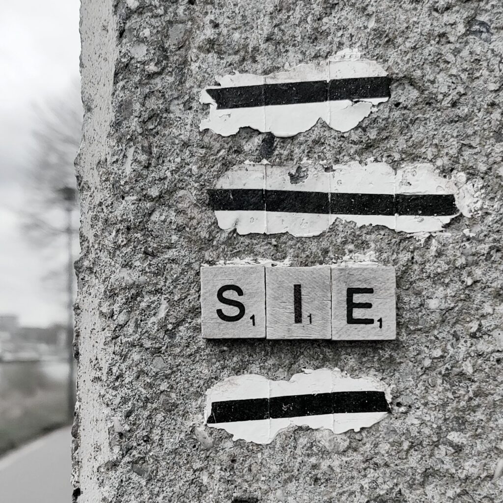A black and white photo of the edge of a wall. Scrabble tiles spelling SIE have two black lines pasted above and one line pasted below.