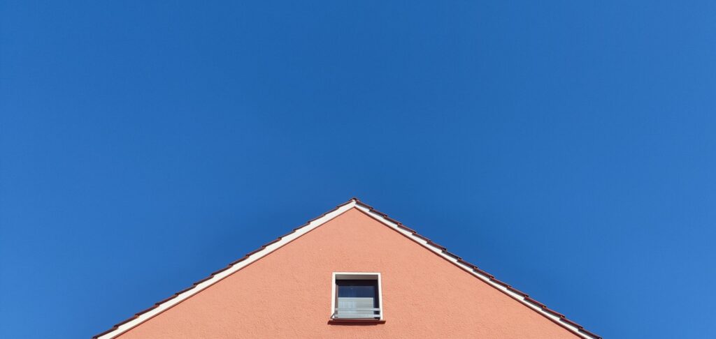 The top of a pink house, with one window, against a bright blue sky.