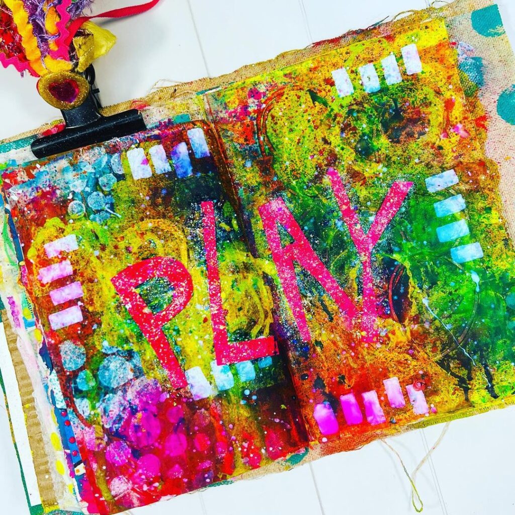 Open mixed media art journal. Multiple colored background with the word PLAY in pink in the middle.