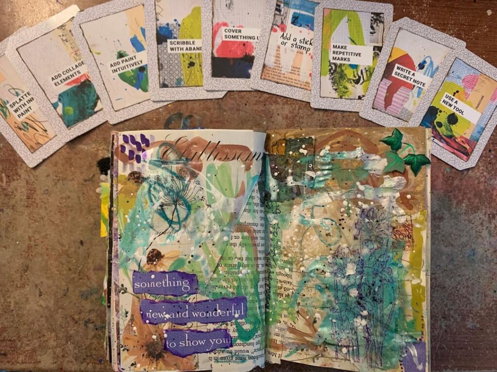 An open mixed media art journal with greens, blues, and browns, and the text "Something new and wonderful to show you" in purple.. Several Ingrid Murray Art Journal Prompt Cards are arranged at the top.