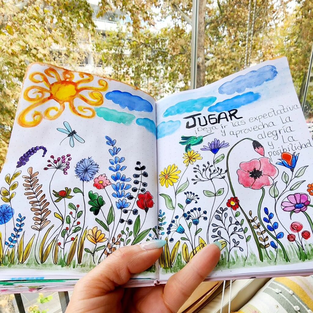 An illustrated art journal page with multi-colored flowers, a sun, and the word "Jugar" (Spanish for "play").