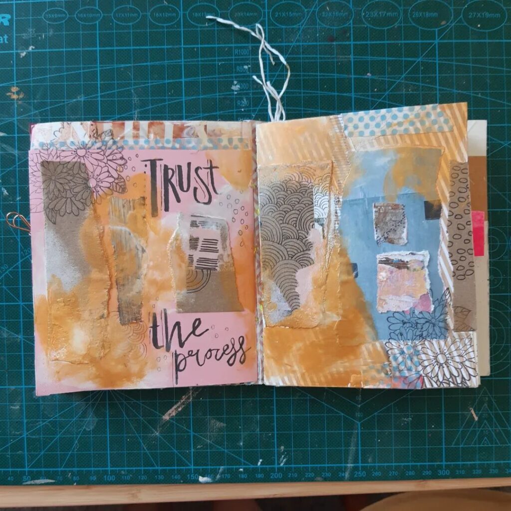 An open mixed media art journal with collage, paint accents in blue, orange, and pink, and script that reads, "Trust the process."
