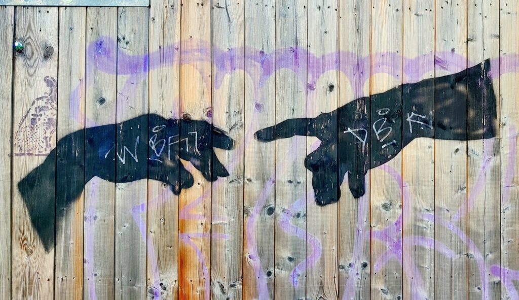 Two outreached hands, spray painted in black onto a wooden fence, extend index fingers as if to touch, a la Michaelangelo's God and Adam on the Sistine Chapel.