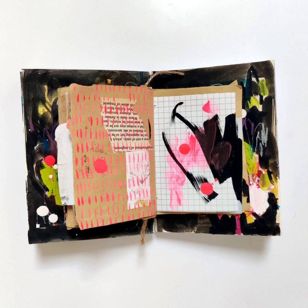Two open mixed media art journals stacked on one another. The one at the bottom has black brush strokes! The one on the top is made of brown craft paper and has minimal collage and energetic marks and drips in neon pink and black.