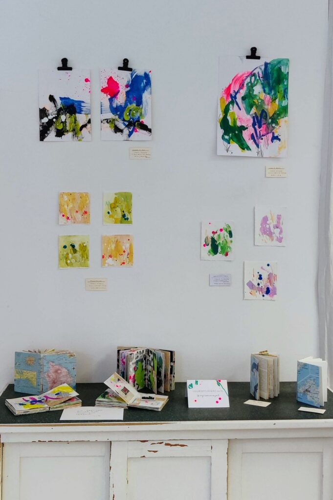 A white wall with 10 colorful abstract art pieces in varying sizes. On a table below sit four completed art journals and two blank art journals.